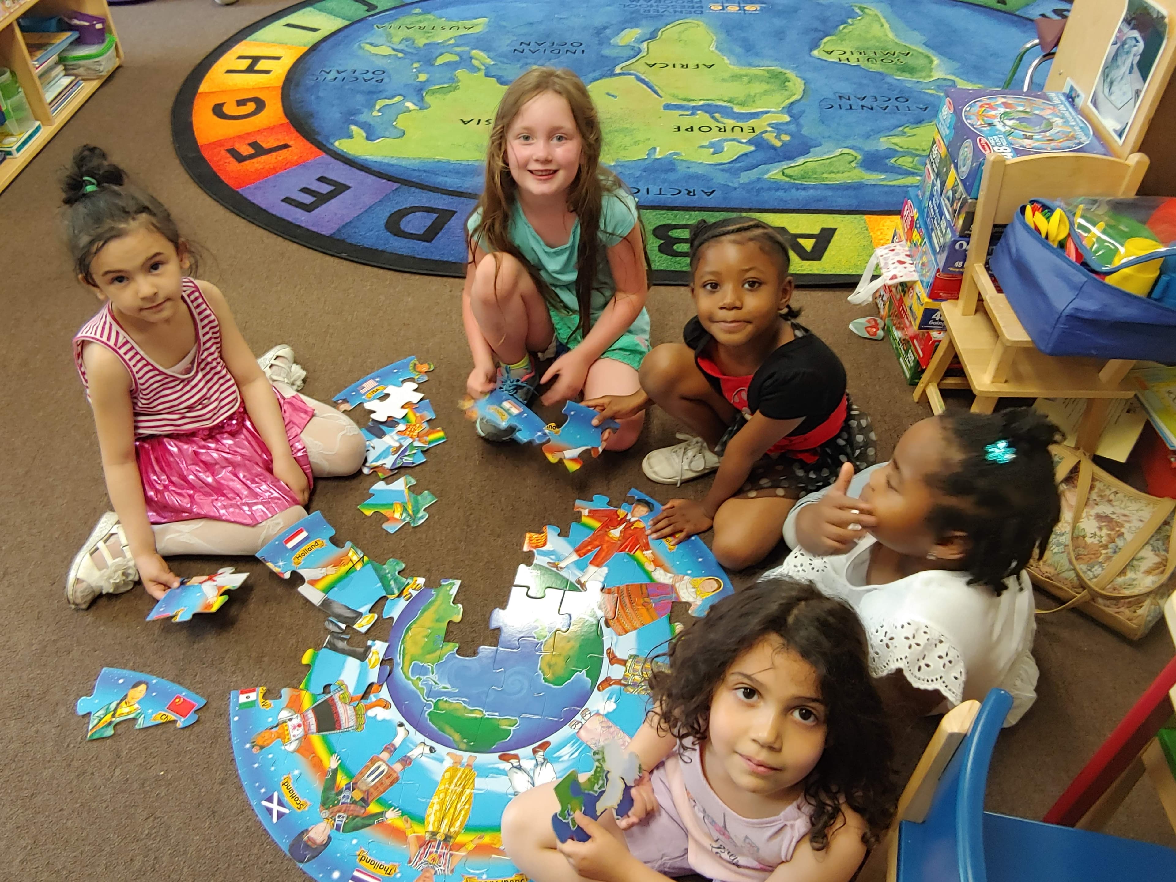 Five preschool girls sit around a giant world puzzle on the floor, smiling up at the camera.