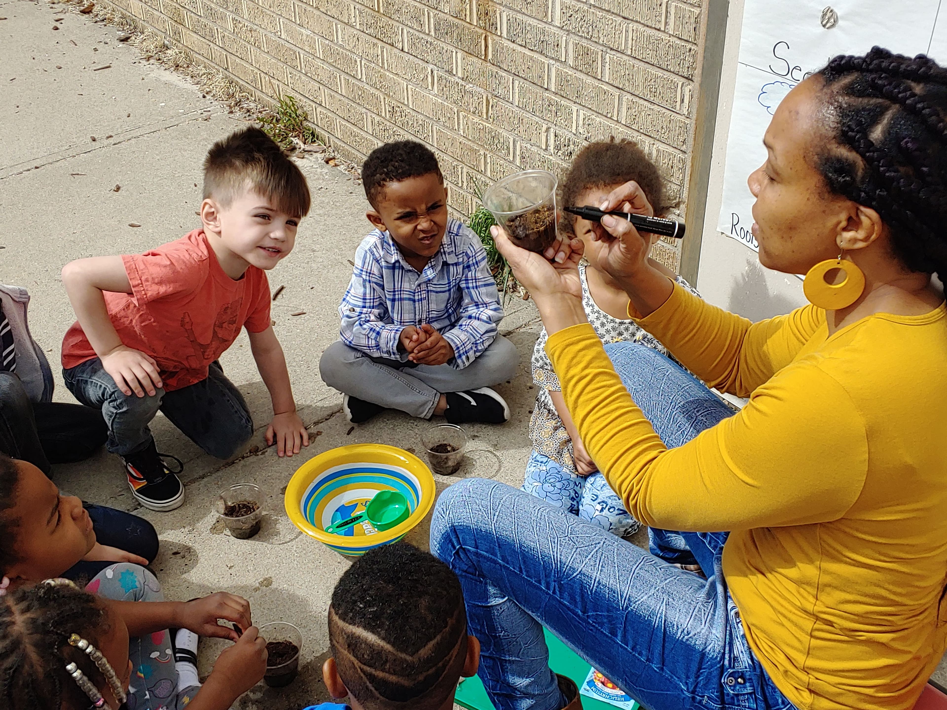 Outside by a brick wall, a teacher holds up a cup of soil while preschoolers watch closely during a planting lesson.