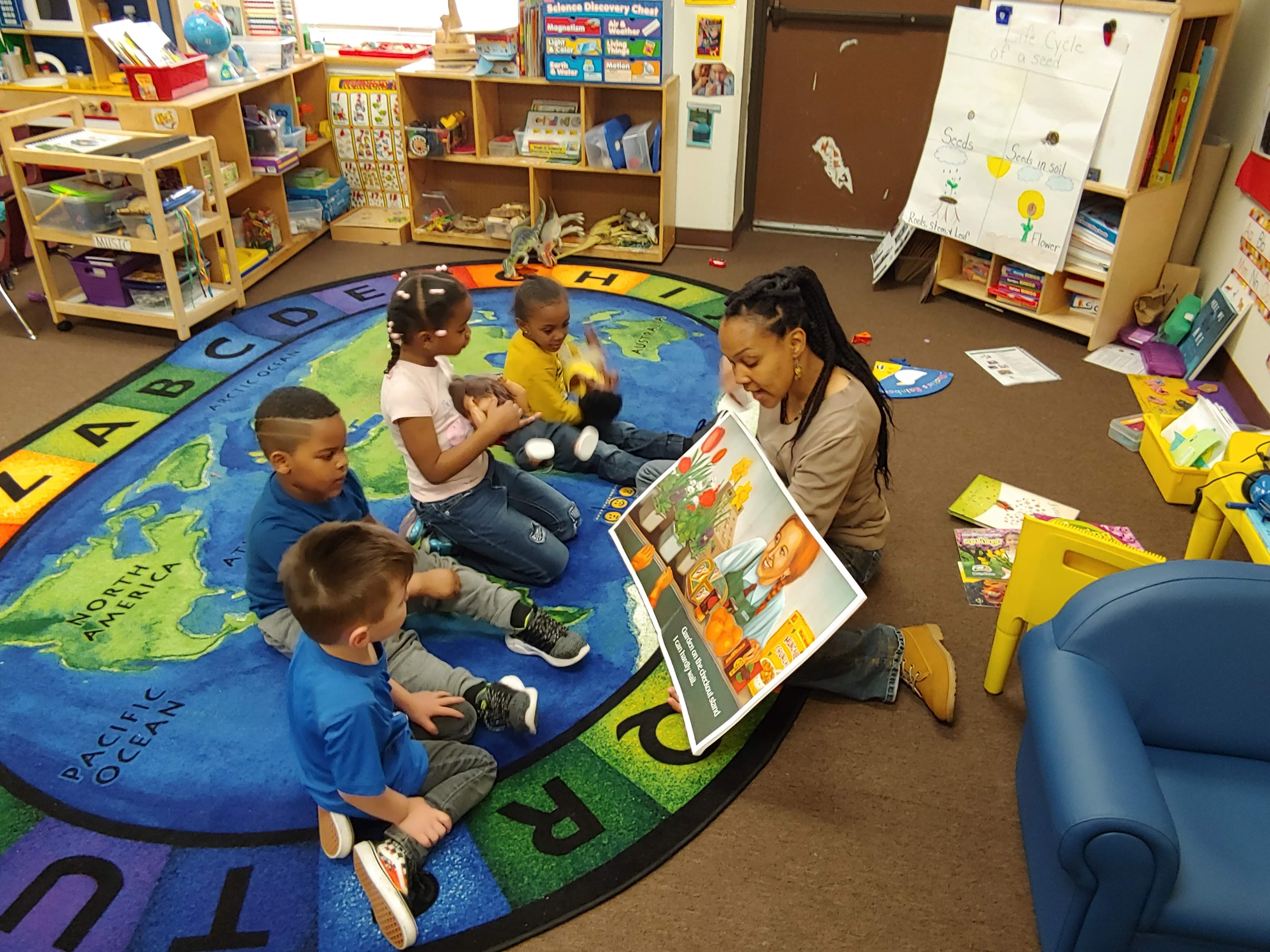 A HOPE Center teacher reads a picture book to four preschoolers on the classroom's alphabet world-map rug.